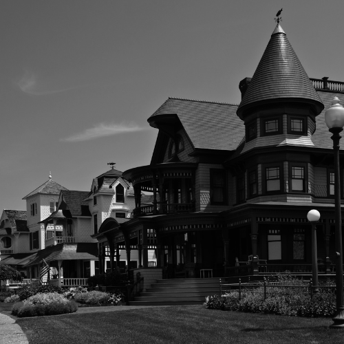 Black and white photo of ornate Victorian houses with a turreted mansion, front porches, landscaped lawn, and street lamps under a clear sky.