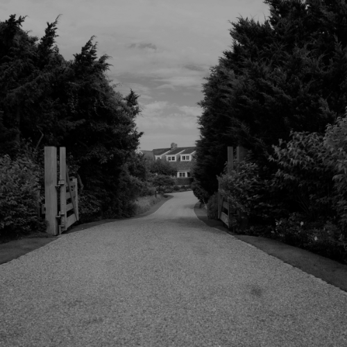 Black and white photo of a gravel driveway lined with tall hedges leading to a large house in the distance.