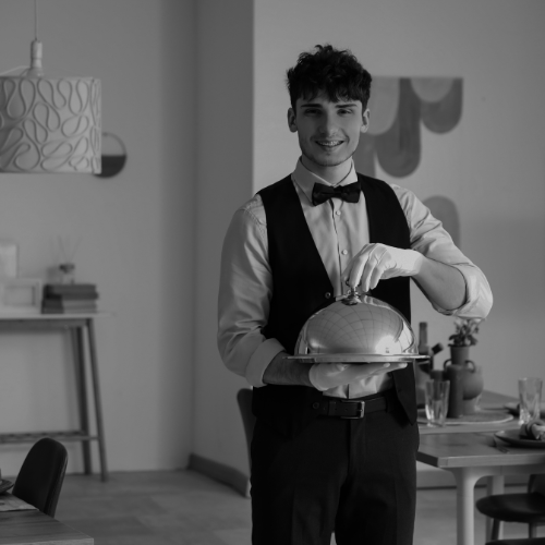 Black and white photo of a smiling server in formal attire holding a covered silver platter inside a restaurant.