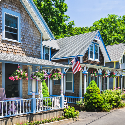Charming seaside cottages with colorful trim, flower baskets, and an American flag along a sunny residential lane.