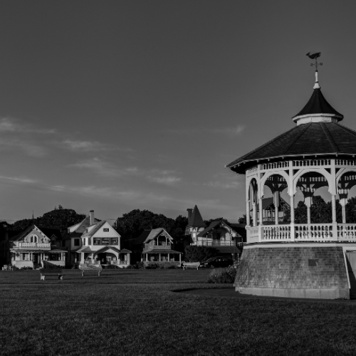 Black and white photo of a large open lawn with a decorative gazebo in the foreground and charming Victorian-style houses in the distance under a wide sky.