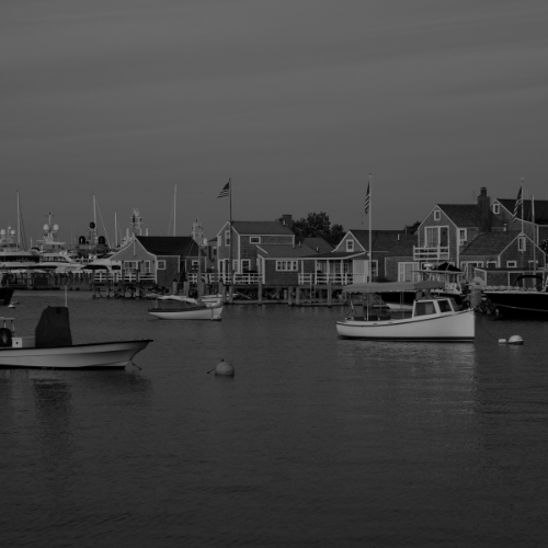 Black and white photo of a harbor with small boats, waterfront cottages, docks, and yachts in the background.