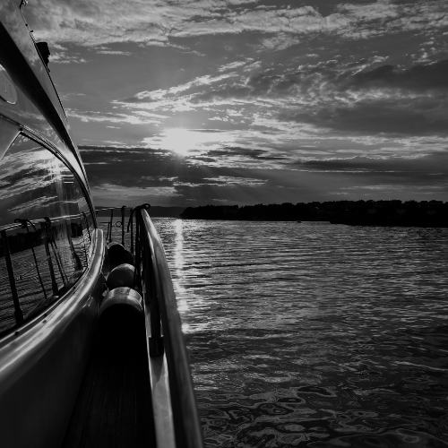 Black and white photo taken from the side of a yacht at sunset, showing calm water and dramatic clouds on the horizon.