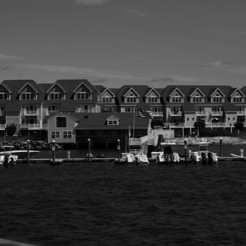 Black and white photo of waterfront townhouses with private docks, boats, and calm harbor water in the foreground.