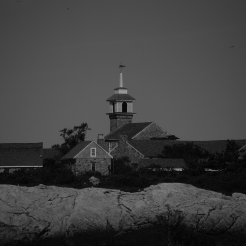 Black and white photo of a stone coastal building with a cupola and weather vane behind rocky shoreline.