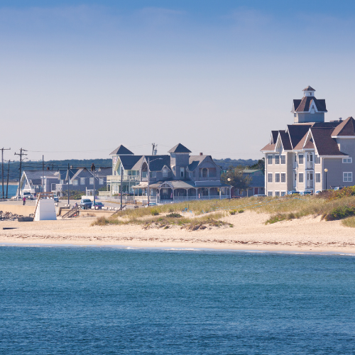 Coastal homes and beachfront buildings along a sandy shoreline with calm blue water in the foreground.