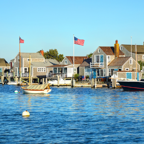 Harborfront cottages and docks with American flags, small boats, and calm blue water under a clear sky.