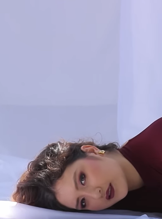 Mujer con cabello rizado, maquillaje de labios oscuros y cabello suelto, recostada en una superficie blanca, vista de perfil