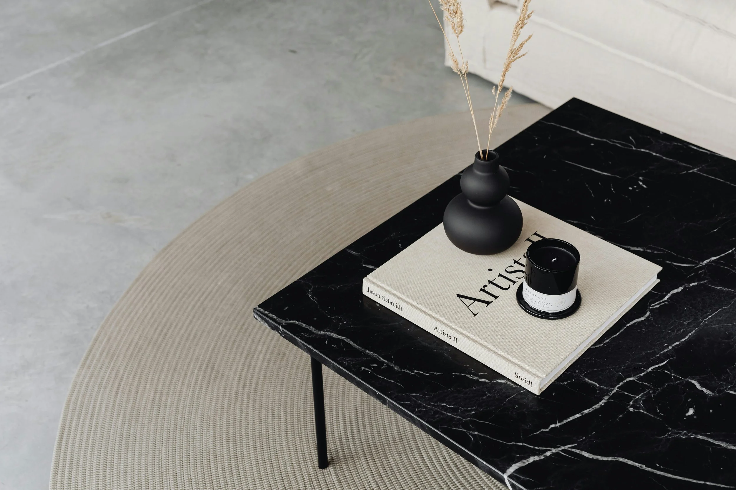 Close-up of a marble top black coffee table with a beige woven rug beneath it, decorated with a black matte vase with dried grass, a hardcover art book titled 'Artistic', and a black candle jar.