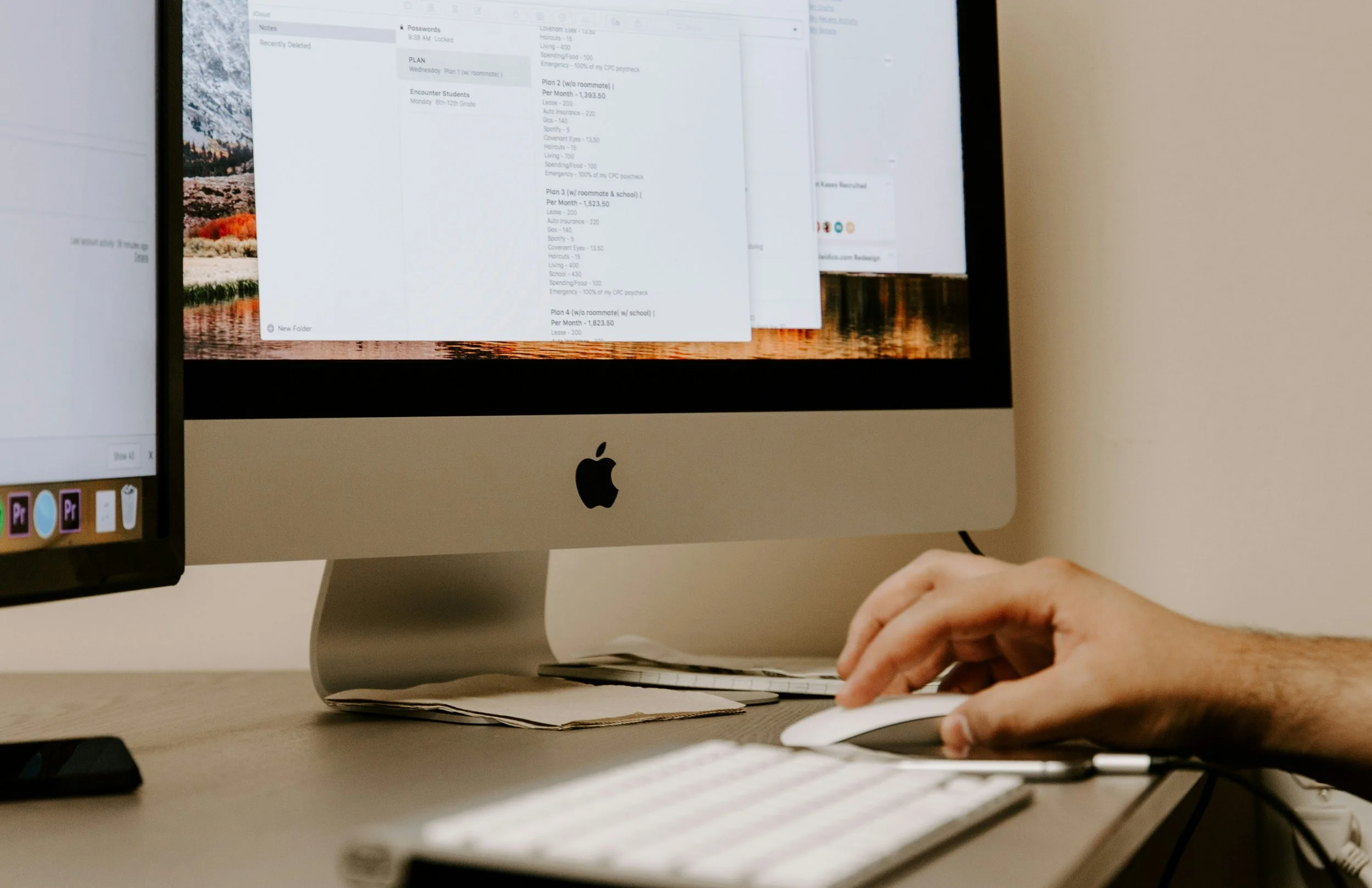 Close-up of a person using a computer mouse and keyboard in front of an Apple iMac with a visible black Apple logo, on a desk with some papers underneath the stand, with a blurred monitor to the left.