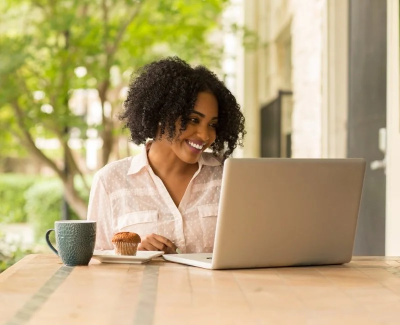 Woman with curly black hair smiling at her laptop at a wooden table outdoors, with a coffee mug and muffin nearby.