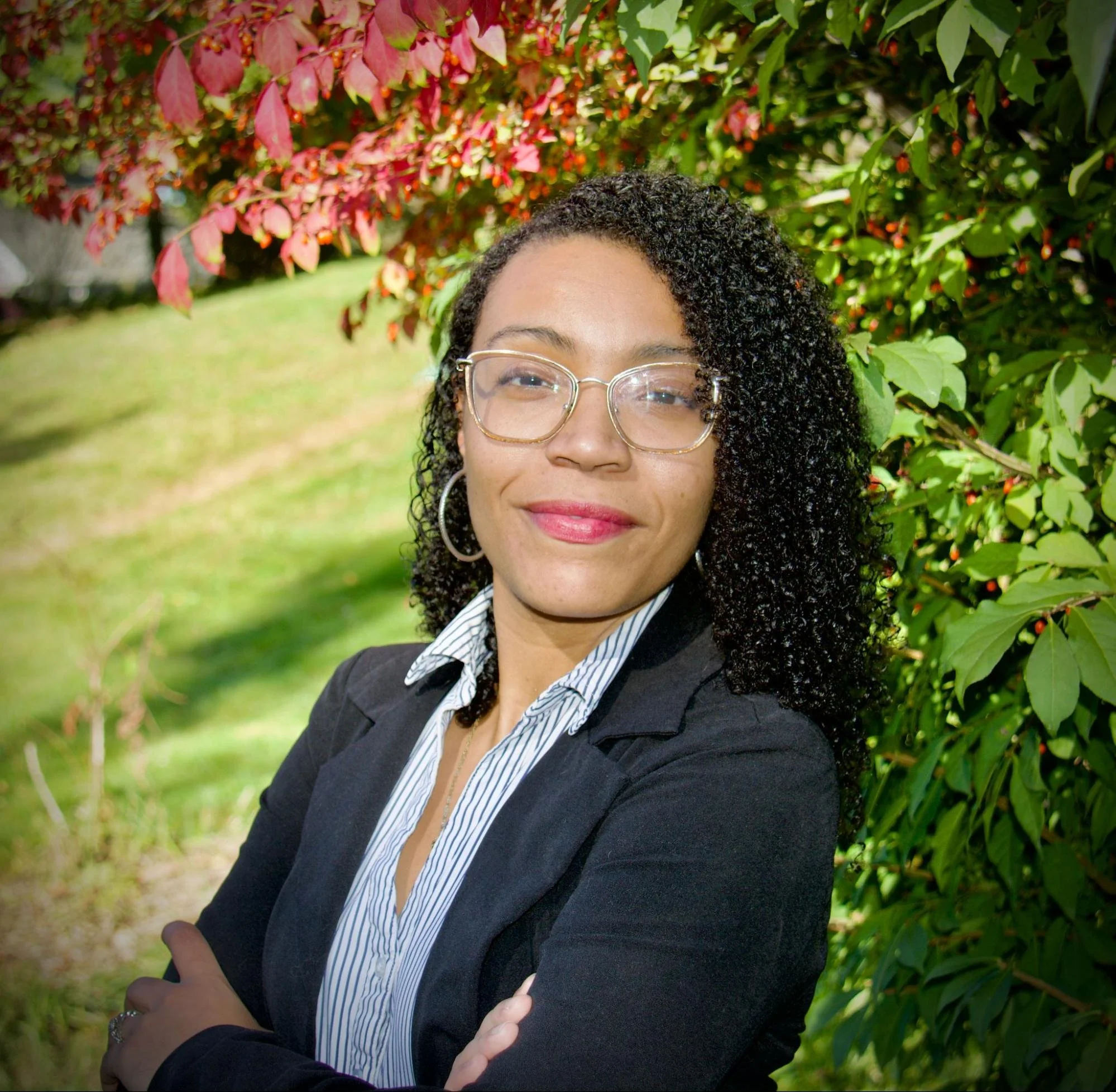 A woman with curly black hair, glasses, and hoop earrings, wearing a black blazer and striped shirt, standing outdoors near green and red foliage.