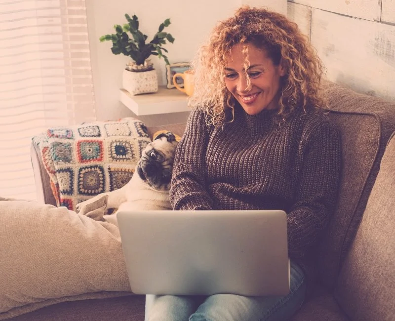 Woman smiling at a laptop while sitting on a couch, with a pug dog resting behind her