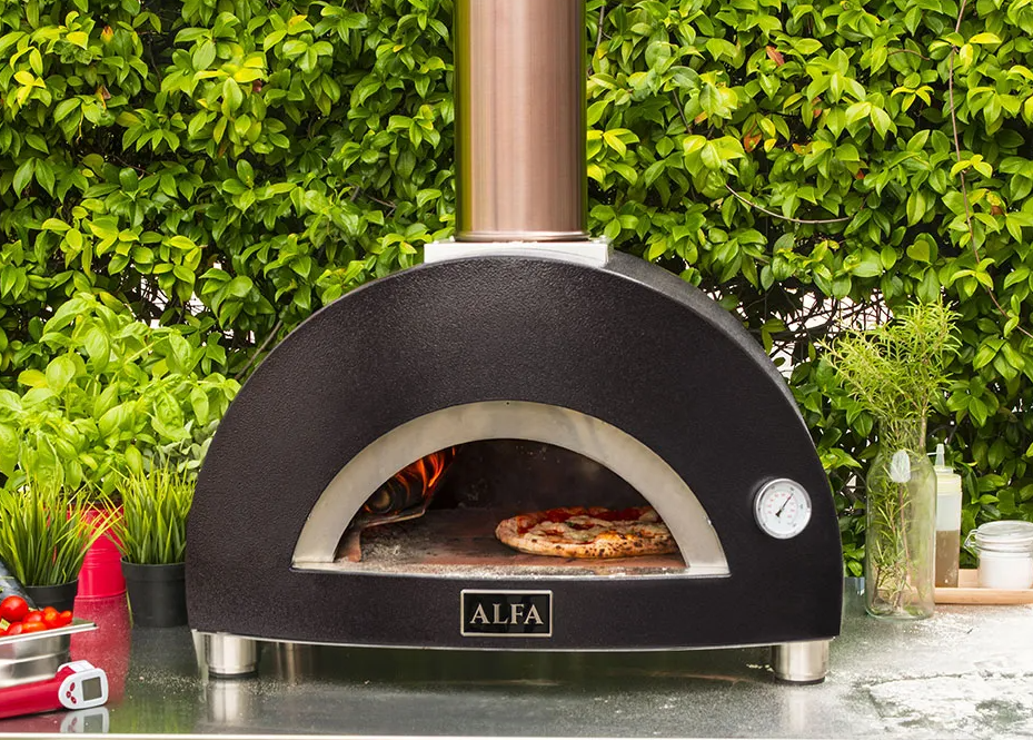 A pizza baking inside a black wood-fired pizza oven outdoors with greenery in the background.
