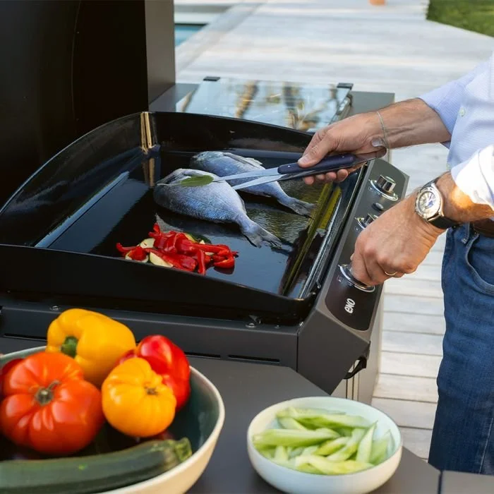 Person grilling fish and red peppers on an outdoor electric grill. Fresh vegetables, including tomatoes, yellow peppers, and cucumber slices, are on the table in front of the grill.