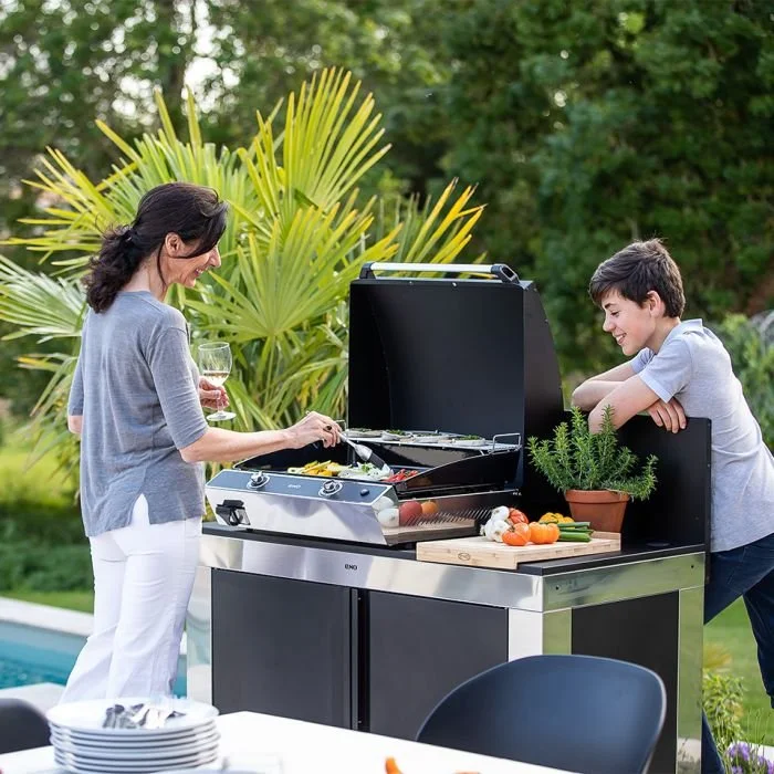 A woman and a boy enjoying a barbecue outdoors, with the woman cooking on a grill and the boy watching, surrounded by greenery and a potted herb plant.
