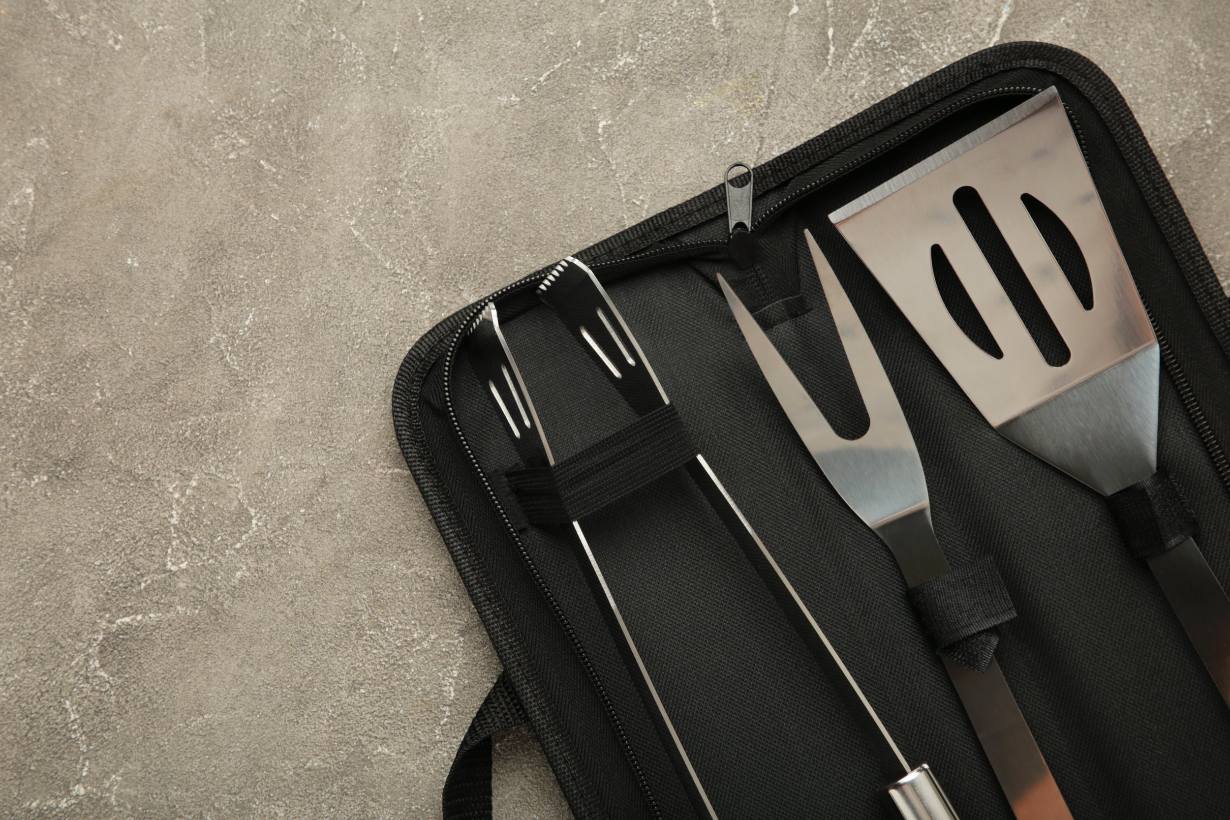 A black zippered case with kitchen utensils including a spatula and a fork inside, on a textured gray surface.