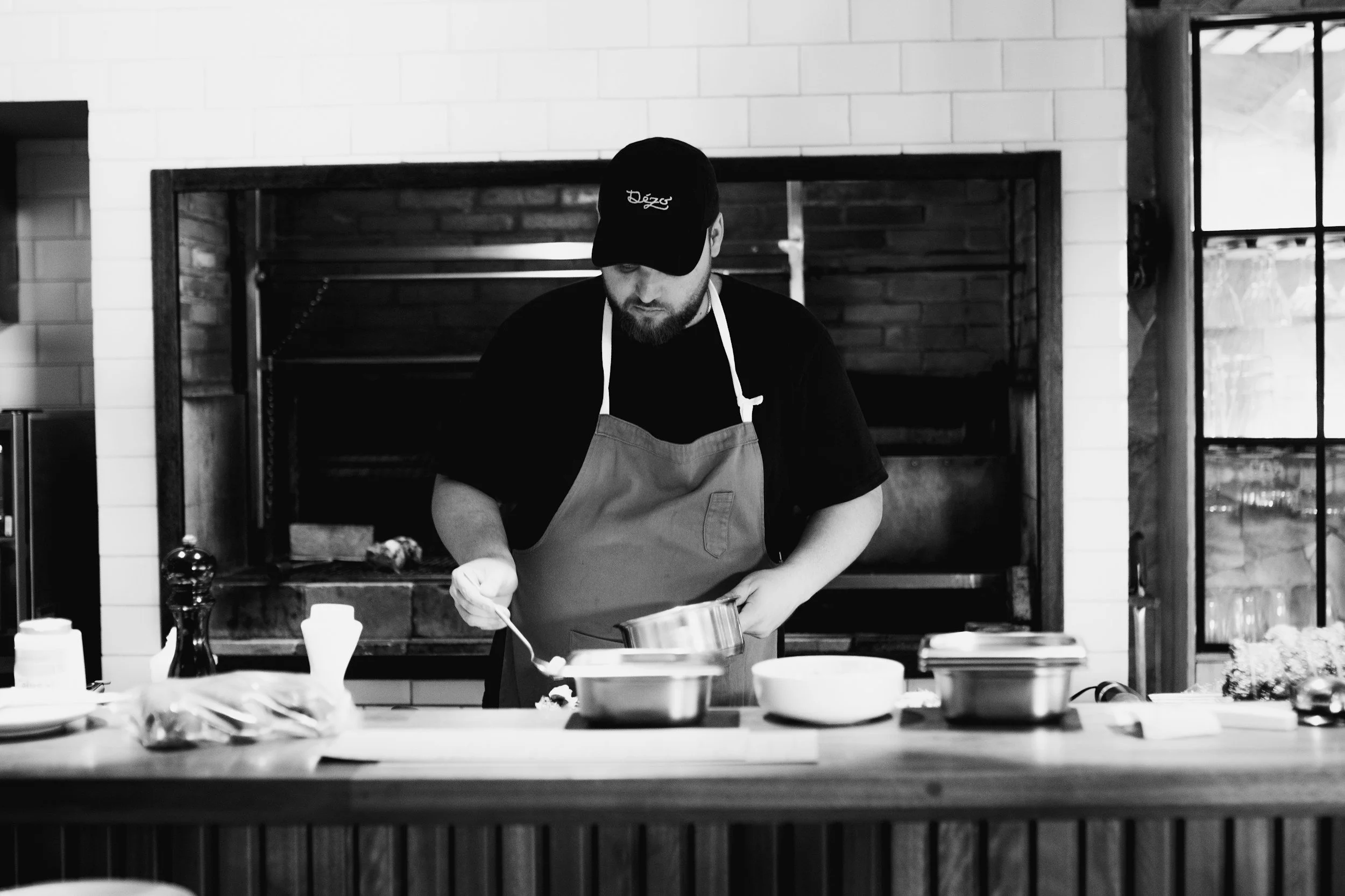 A man in a baseball cap, black t-shirt, and apron preparing food at a kitchen counter in front of a brick oven, in black and white.