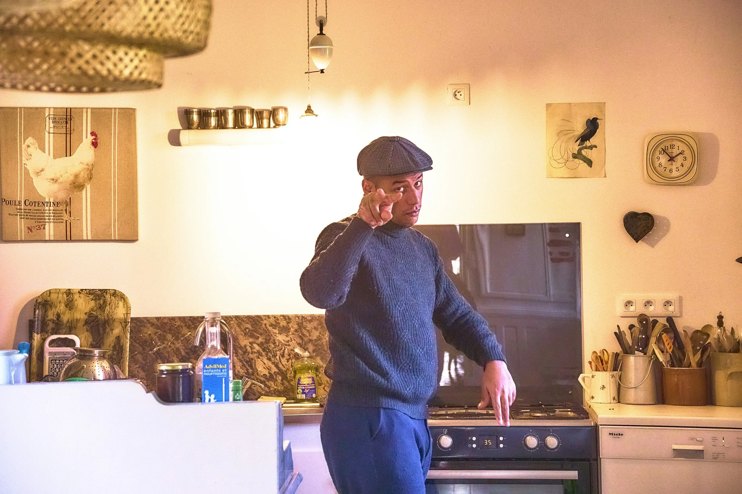 A man wearing a flat cap and navy sweater standing in a kitchen, pointing at the camera with a serious expression, behind a stove and kitchen utensils.