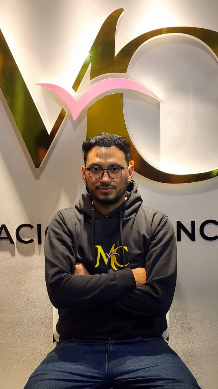 Joven con gafas, camisa con logotipo MCA, sentado en una silla, fondo con logotipo y letras.