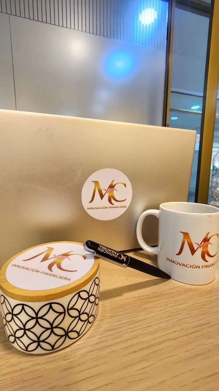 Varios objetos sobre una mesa de madera en un entorno interior, incluyendo una taza blanca, un bol con diseño negro y tapa de madera, un bolígrafo negro, y una laptop plateada con un logotipo de "Innovación Financiera" en el centro, así como en la ta