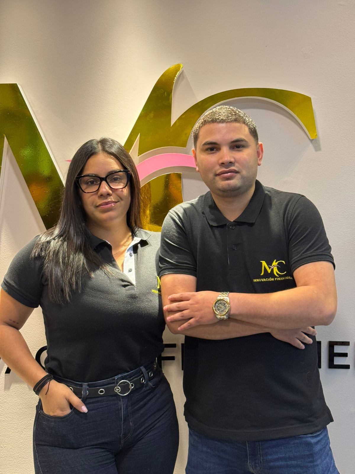 Dos personas, un hombre y una mujer, posando frente a un logotipo en una oficina, ambos con ropa negra con logotipo y el fondo de un muro blanco.