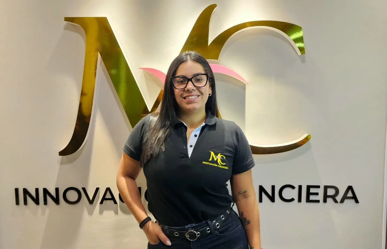 Mujer con gafas y camiseta negra con logotipo de NC, sonriendo frente a un logo dorado y rosa en una pared