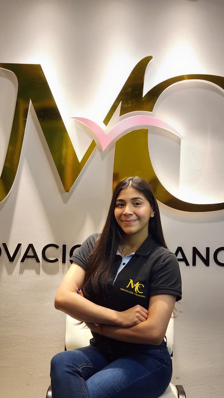Joven sentada en una sala de oficina, viste una camiseta de color negro con logotipo y letras amarillas, y sonríe en una pose con los brazos cruzados.