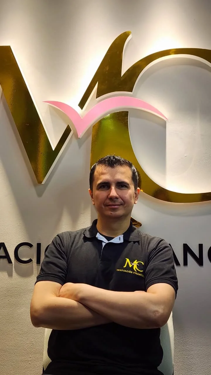 Hombre con camiseta negra con logotipo amarillo 'MC Innovación Financiera' posando con brazos cruzados frente a un logo grande en la pared.