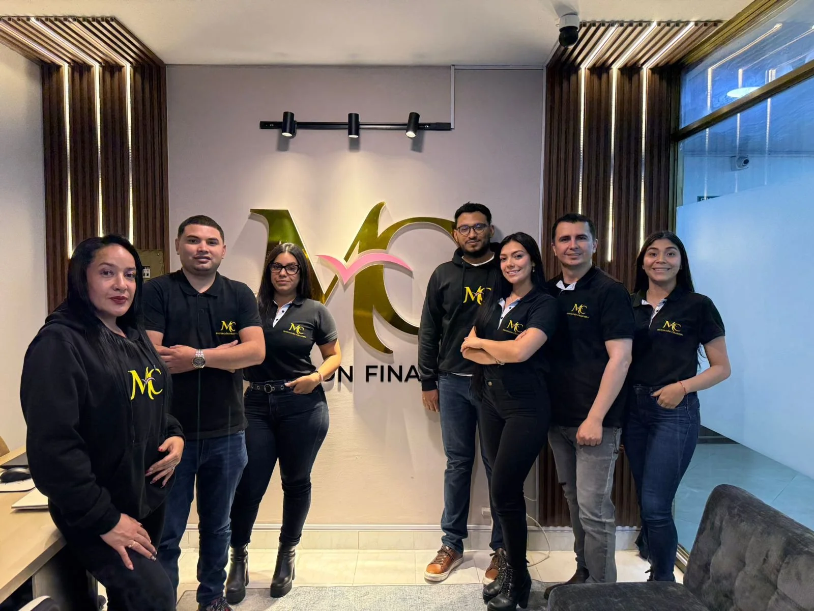 Grupo de siete personas posando en una oficina con un logo en la pared de fondo que tiene letras doradas y rosas. Todos llevan camisetas o sudaderas negras con un logotipo amarillo que dice MC.
