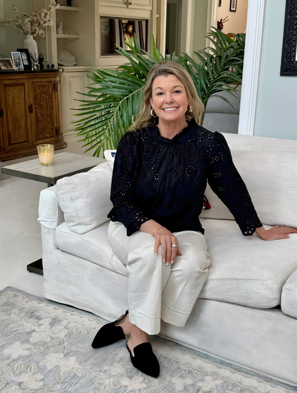 A woman sitting on a white sofa in a living room, smiling at the camera, with a large green plant behind her, and a wooden cabinet with framed photos and decor on the left side.