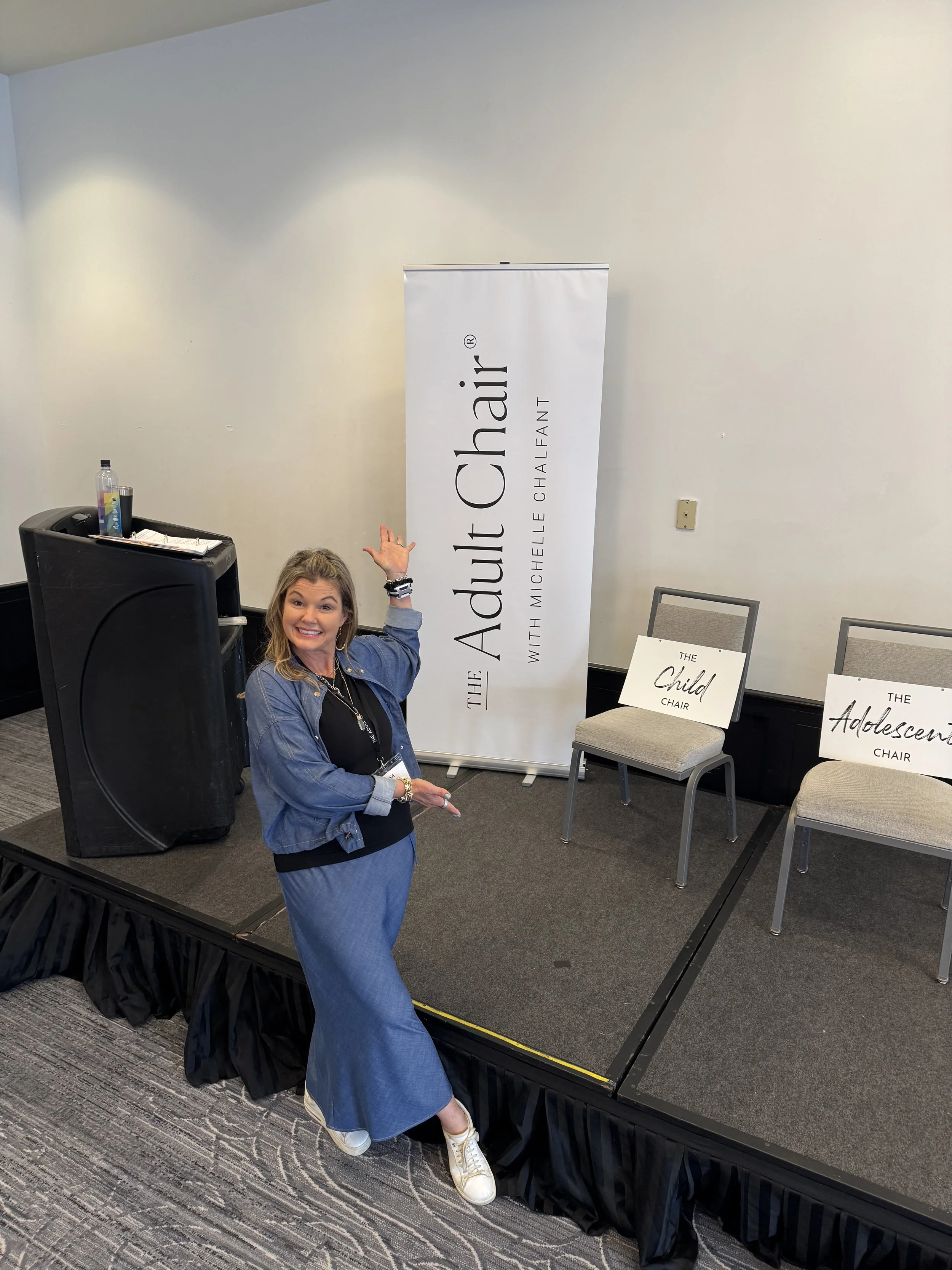 A woman standing on a stage next to a music speaker and a large vertical banner that reads 'The Adult Chair with Michelle Chalfaint.' She is smiling, pointing upward with her right hand, and gesturing with her left hand. Two chairs on the stage have signs that say 'The Child Chair' and 'The Adolescent Chair.'