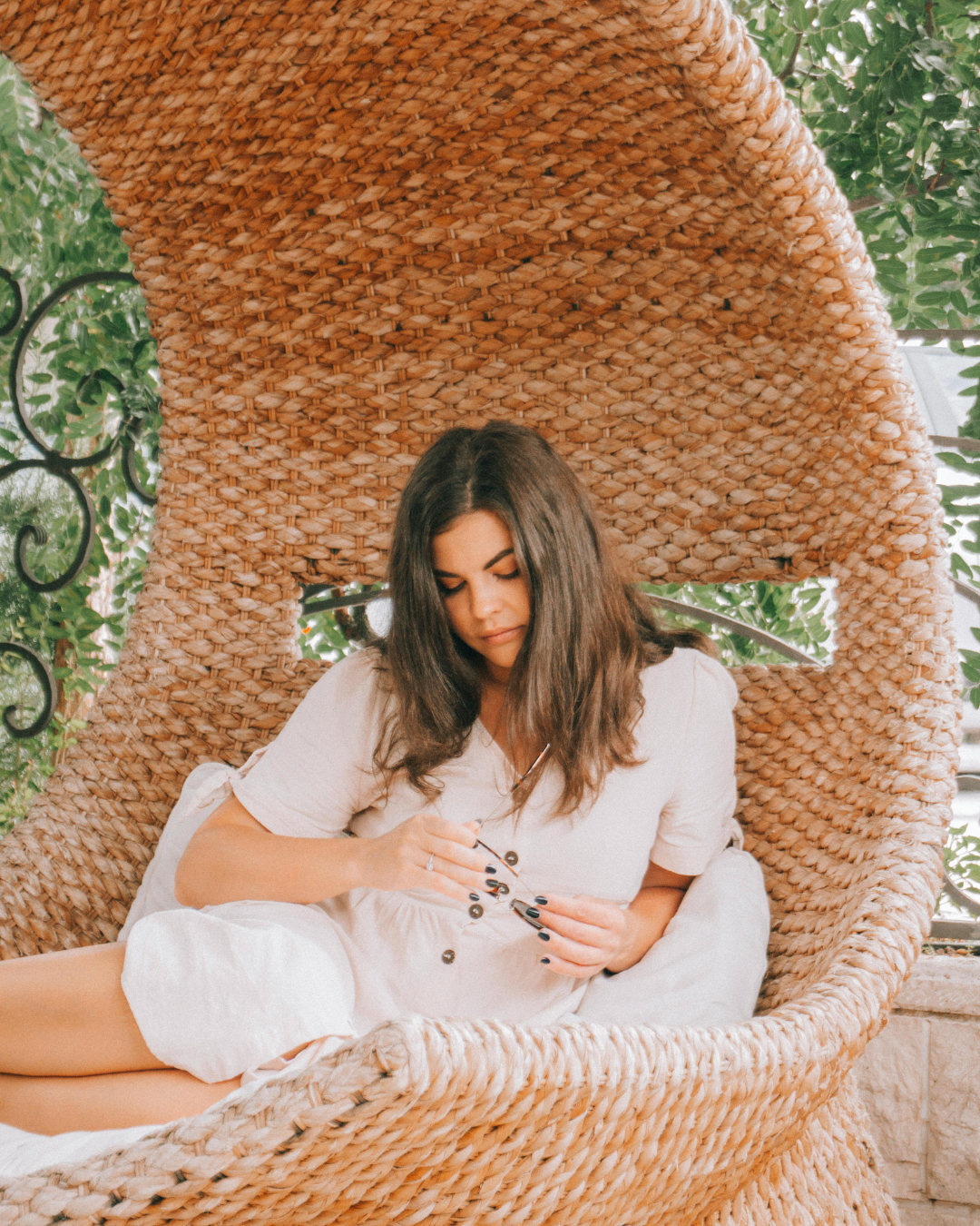 A young woman with dark hair sitting in a large woven wicker chair outdoors, surrounded by green foliage, wearing a light-colored dress and holding a small object.