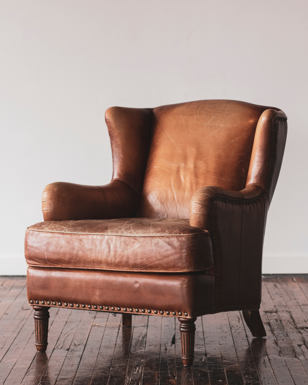 A vintage brown leather armchair with wooden legs positioned on a hardwood floor, against a plain light-colored wall.