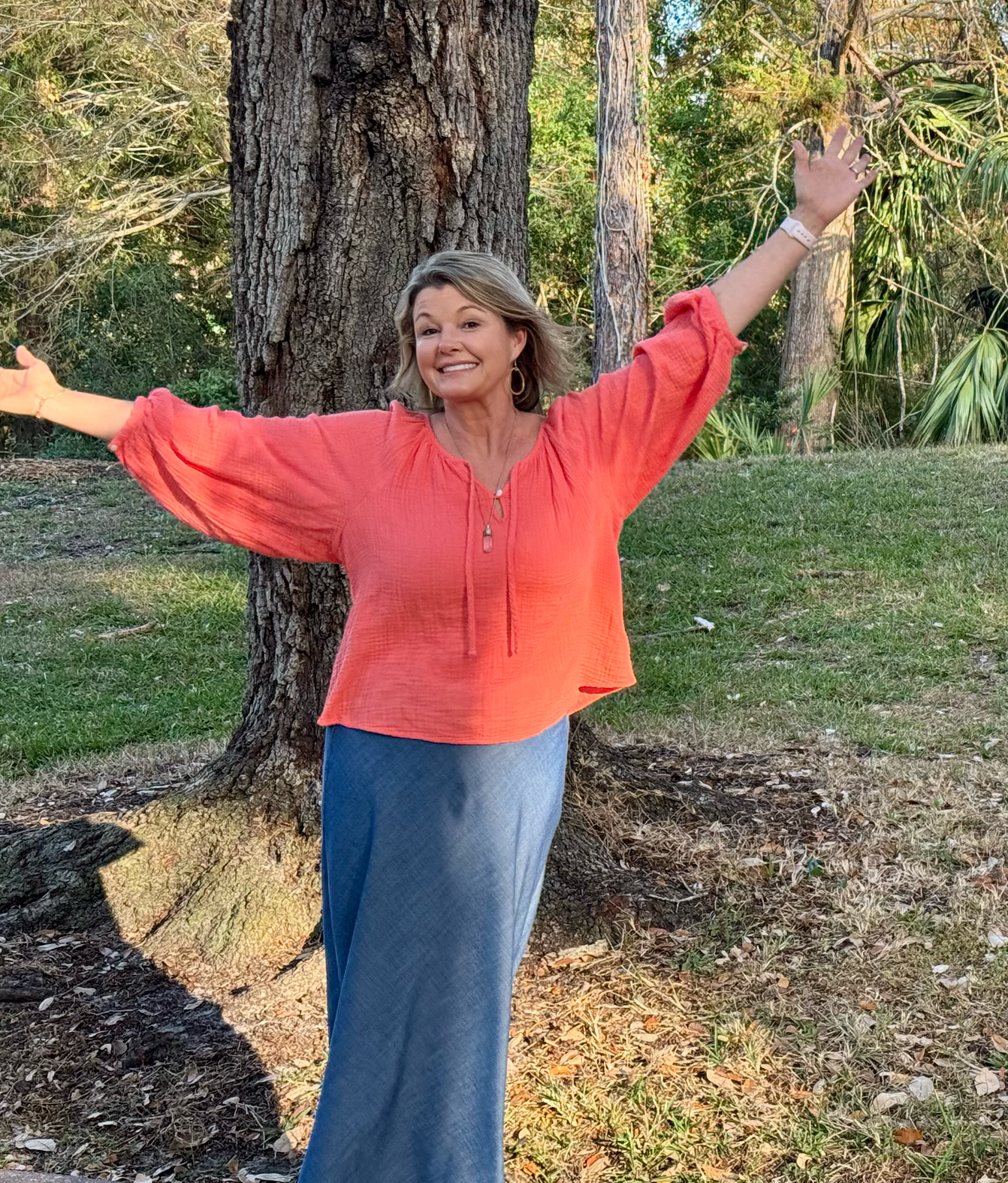 Woman in a coral long-sleeve blouse with arms outstretched outdoors near a large tree.