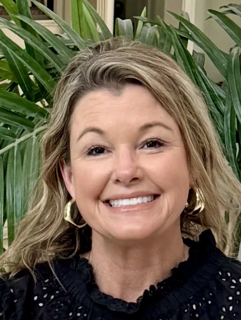 A woman with blond wavy hair smiling, wearing gold hoop earrings and a black top with eyelet details, standing in front of green leafy plants.