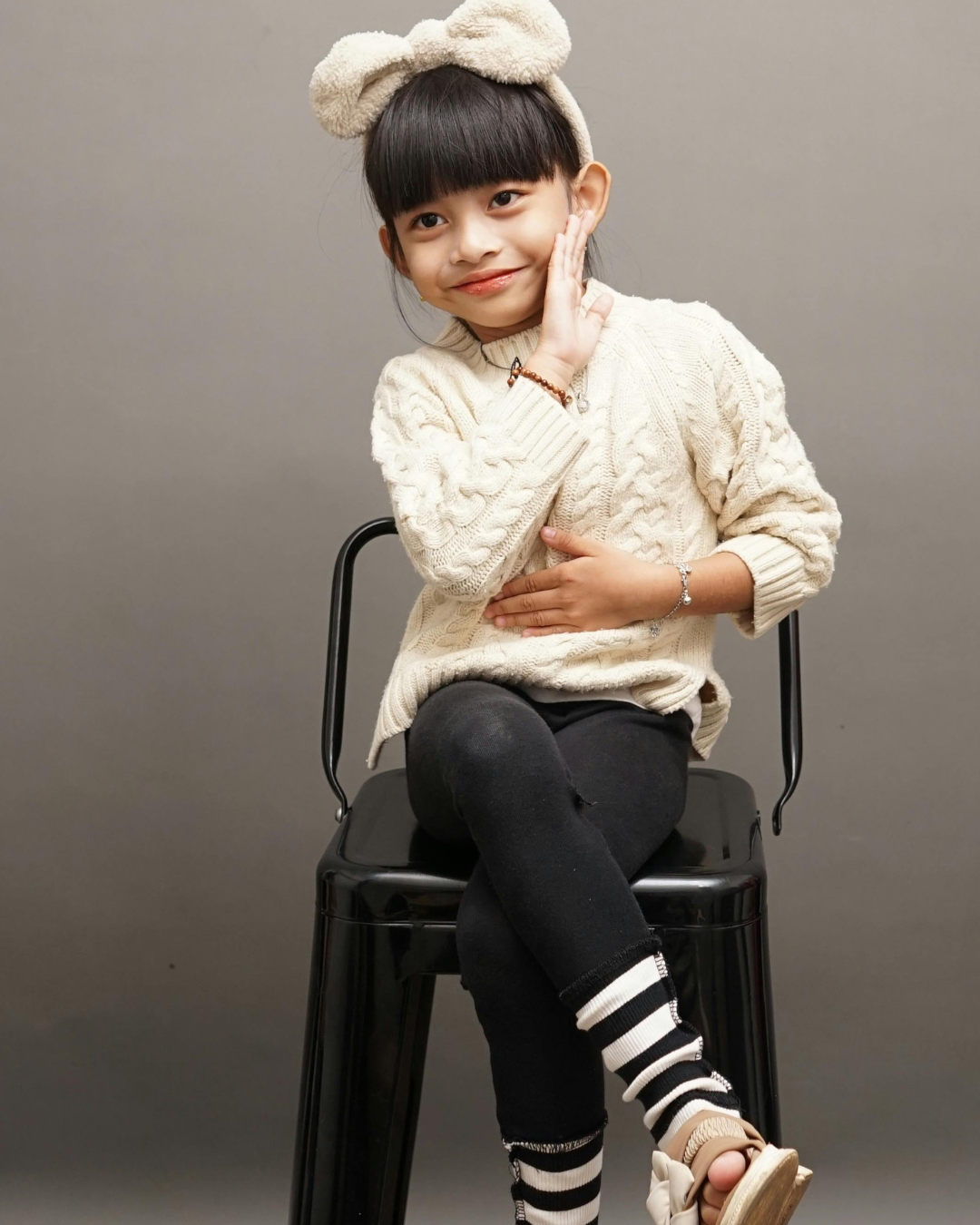 Young girl sitting on a black chair with her legs crossed, wearing a cream-colored sweater, black pants, and striped socks. She has a fluffy headband with bear ears and is touching her cheek with one hand while resting her other hand on her stomach, smiling softly against a gray background.