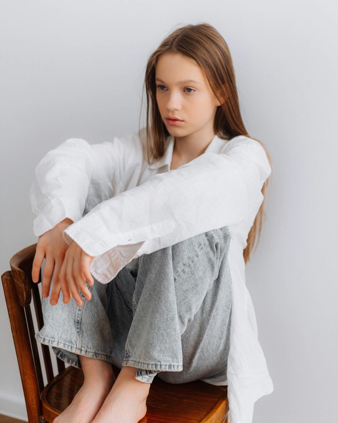 A young woman with long red hair sitting on a wooden chair, wearing a white shirt and gray jeans, with a neutral expression against a plain white wall.