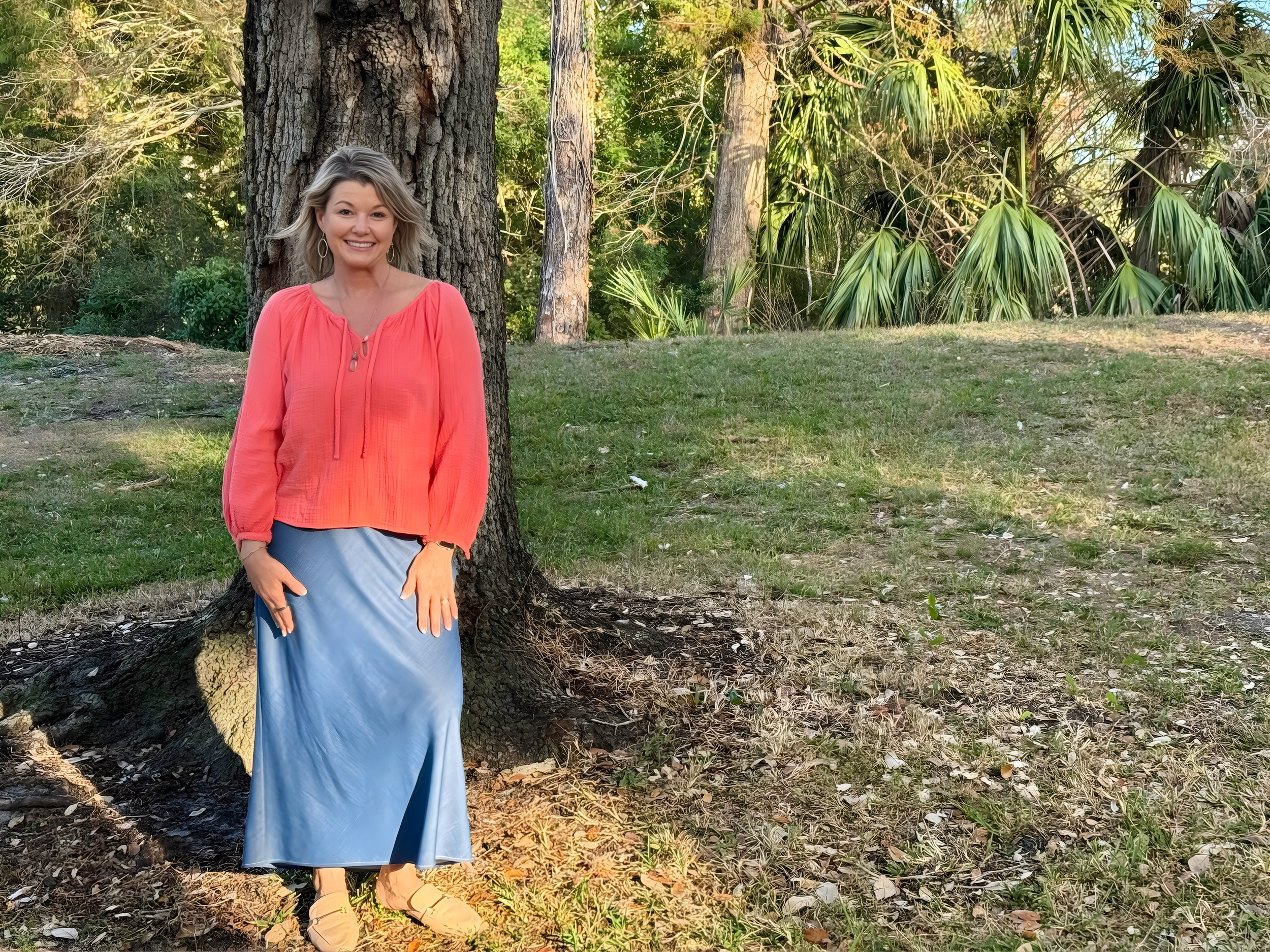 A woman standing outdoors next to a tree in a grassy area surrounded by trees and foliage, wearing a coral long-sleeve top and a long blue skirt.