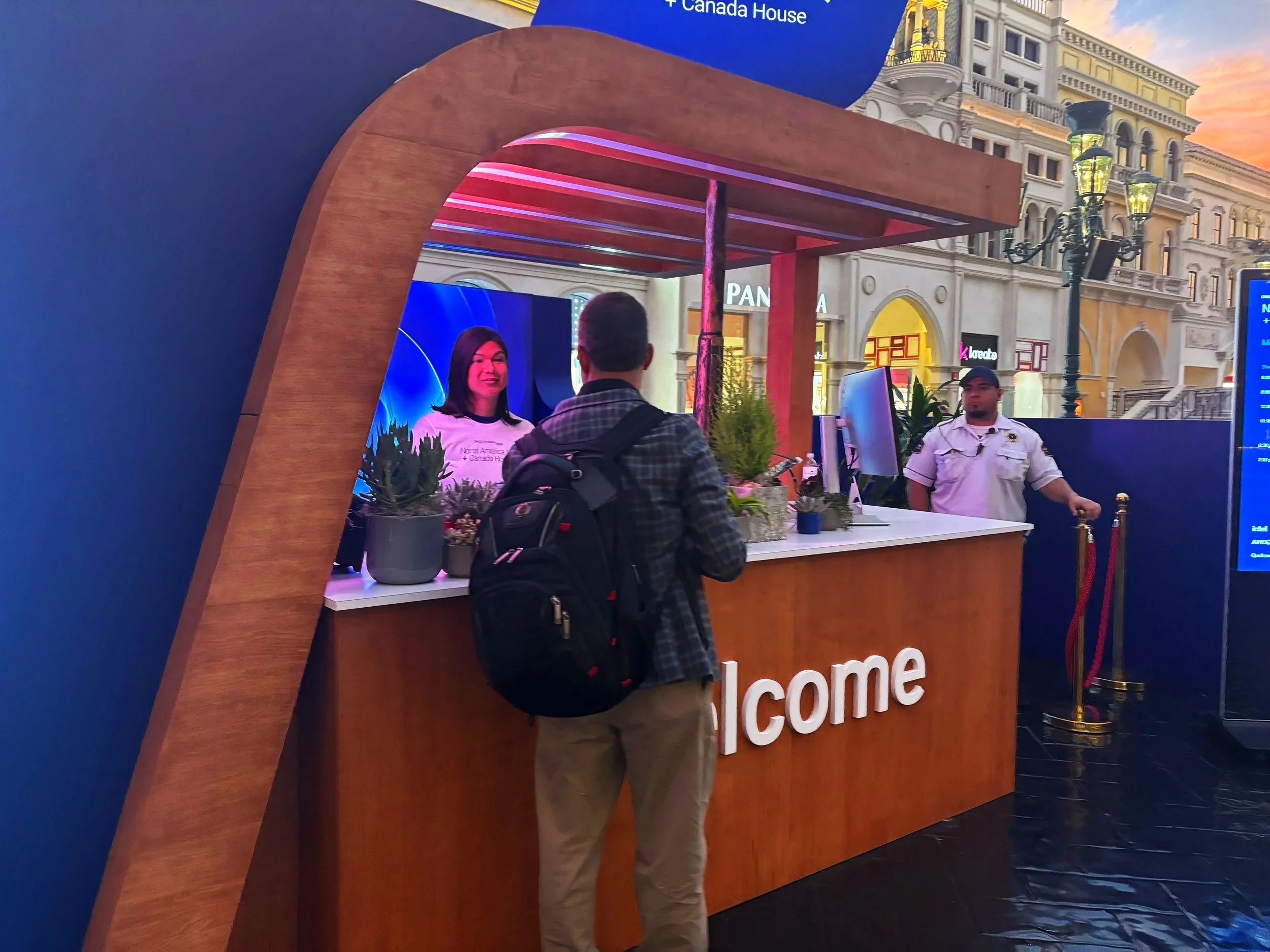 A man with a backpack at a reception desk speaking to a woman behind the counter, which has a large 'Welcome' sign, in an indoor setting with a cityscape backdrop.
