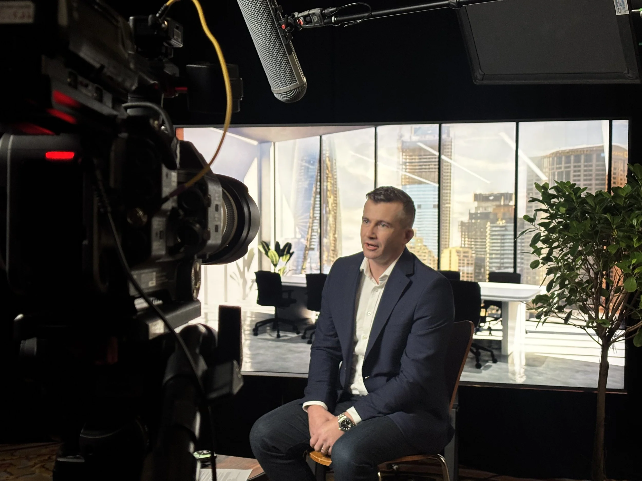 A man in a dark suit sitting in front of a camera during an interview or filming session in a modern office with large windows and a city skyline in the background.