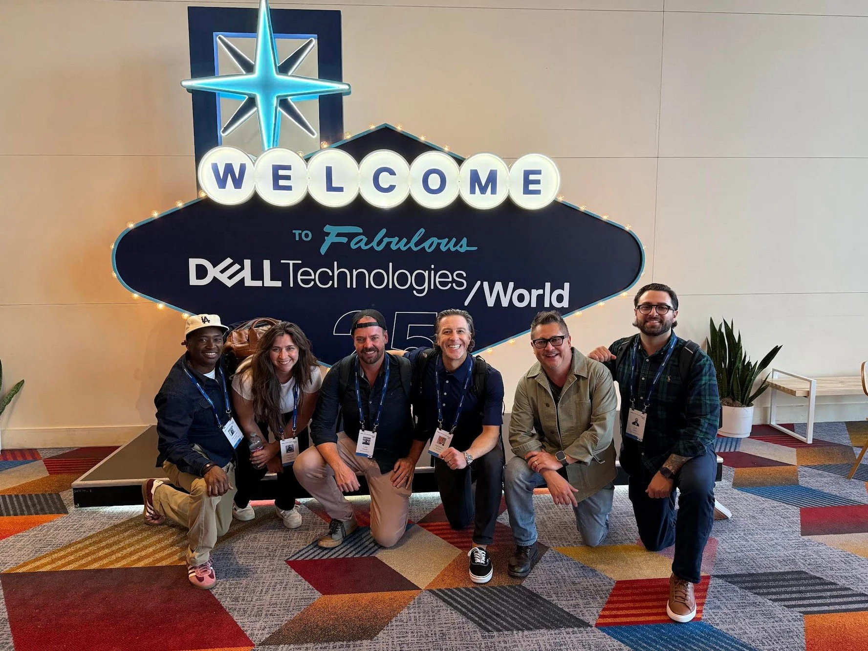 Group of six people kneeling in front of a sign that reads "Welcome to Fabulous Dell Technologies World." They are smiling and wearing conference badges.
