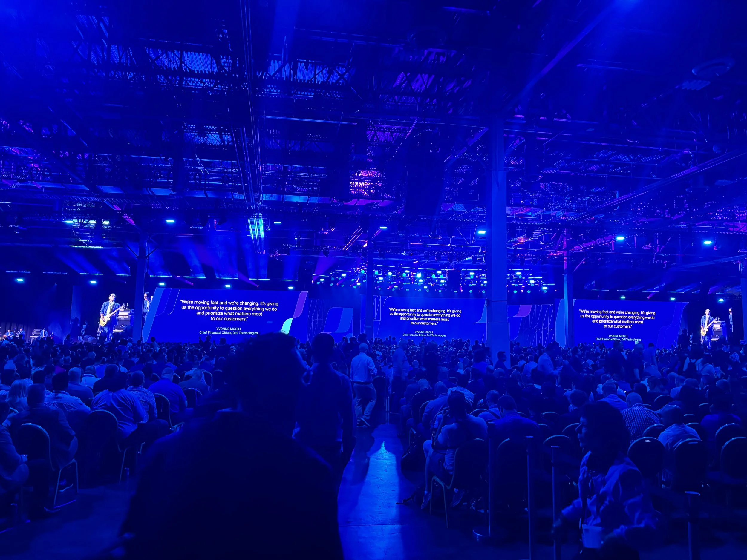 Large indoor conference with many attendees seated and a stage with a band playing, large screens displaying text, and vibrant blue lighting overhead.