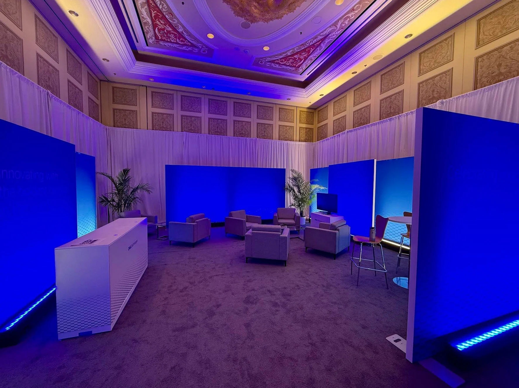 An indoor conference room with purple lighting, beige carpet, patterned ceiling, and blue display boards forming a semi-circle. There are beige armchairs arranged in a circle, a flat-screen TV, potted plants, and a white counter.