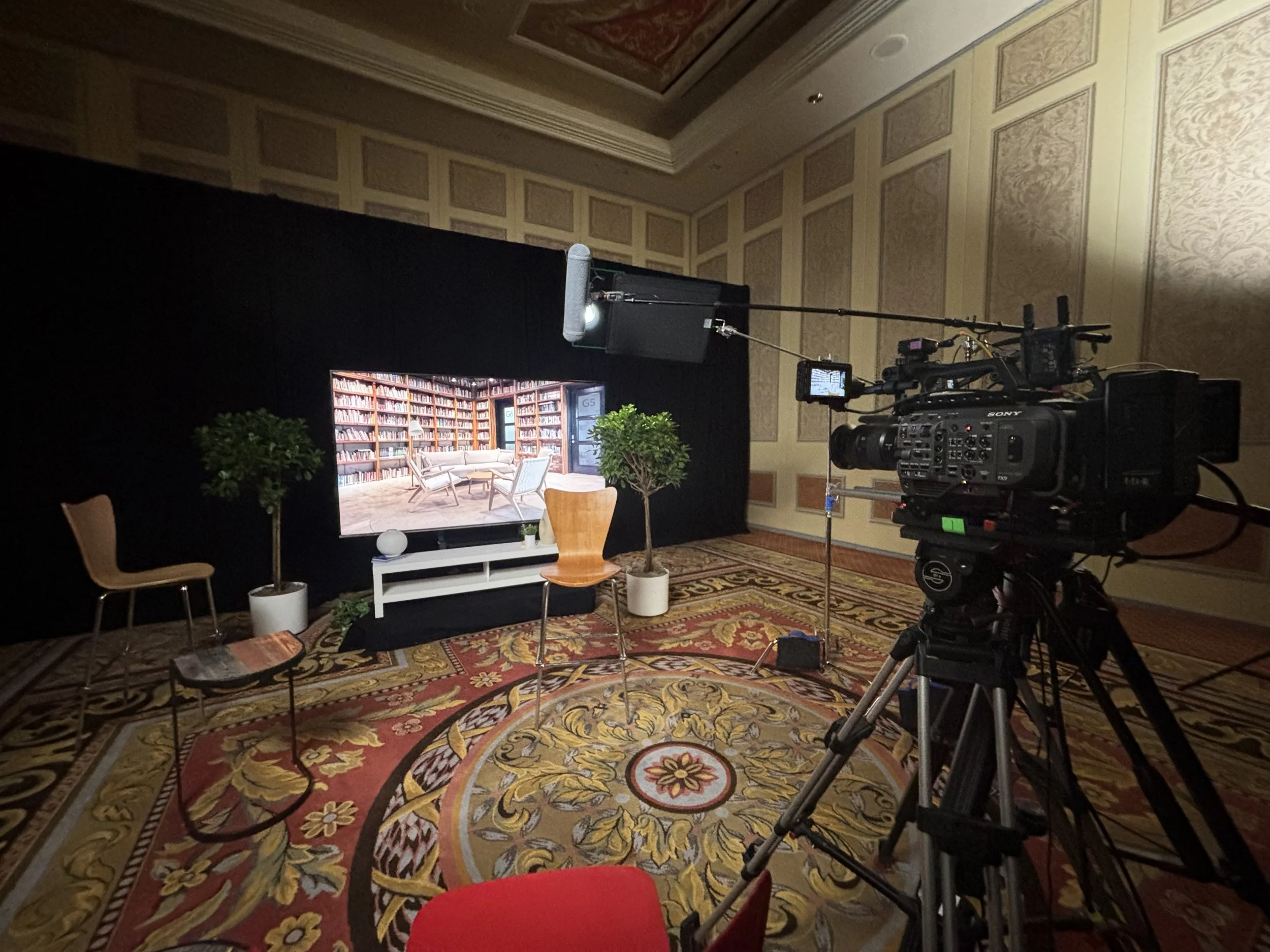 A recording studio setup with a professional camera on a tripod, a large monitor displaying a room with bookshelves, chairs, and plants, and a backdrop with chairs and plants on a decorative carpeted floor.