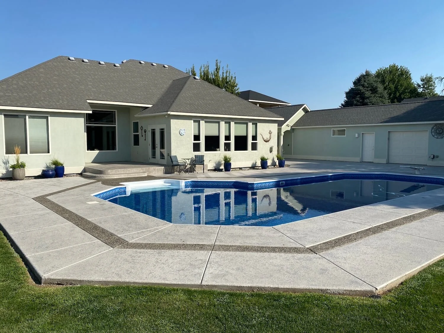 Backyard view of a house with a swimming pool and patio. The house has beige siding, multiple windows, and a gray roof. There are potted plants, patio furniture, and decorative items around the pool area, with a clear blue sky overhead.