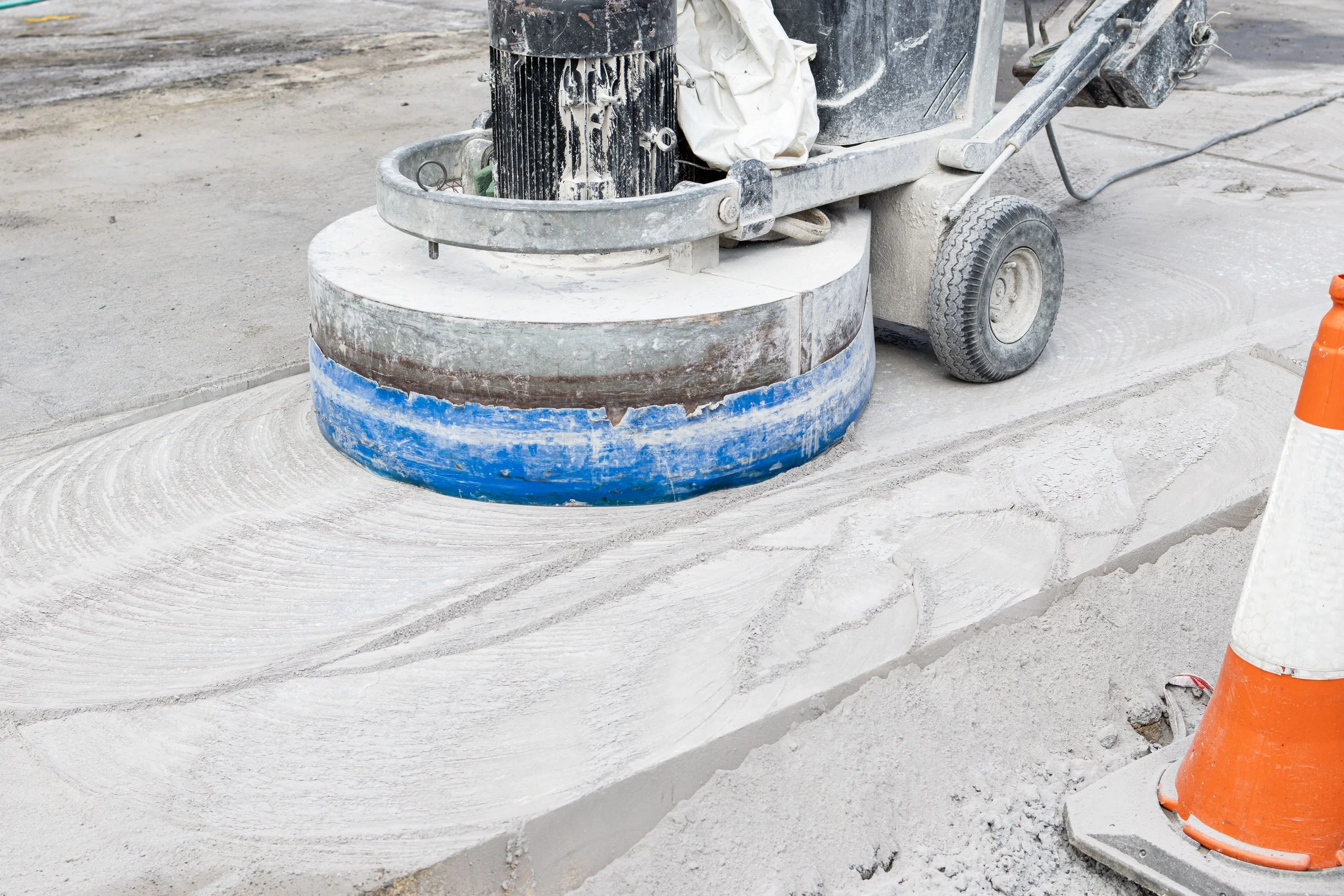 Concrete surface being polished with a floor grinder, with orange safety cone nearby.