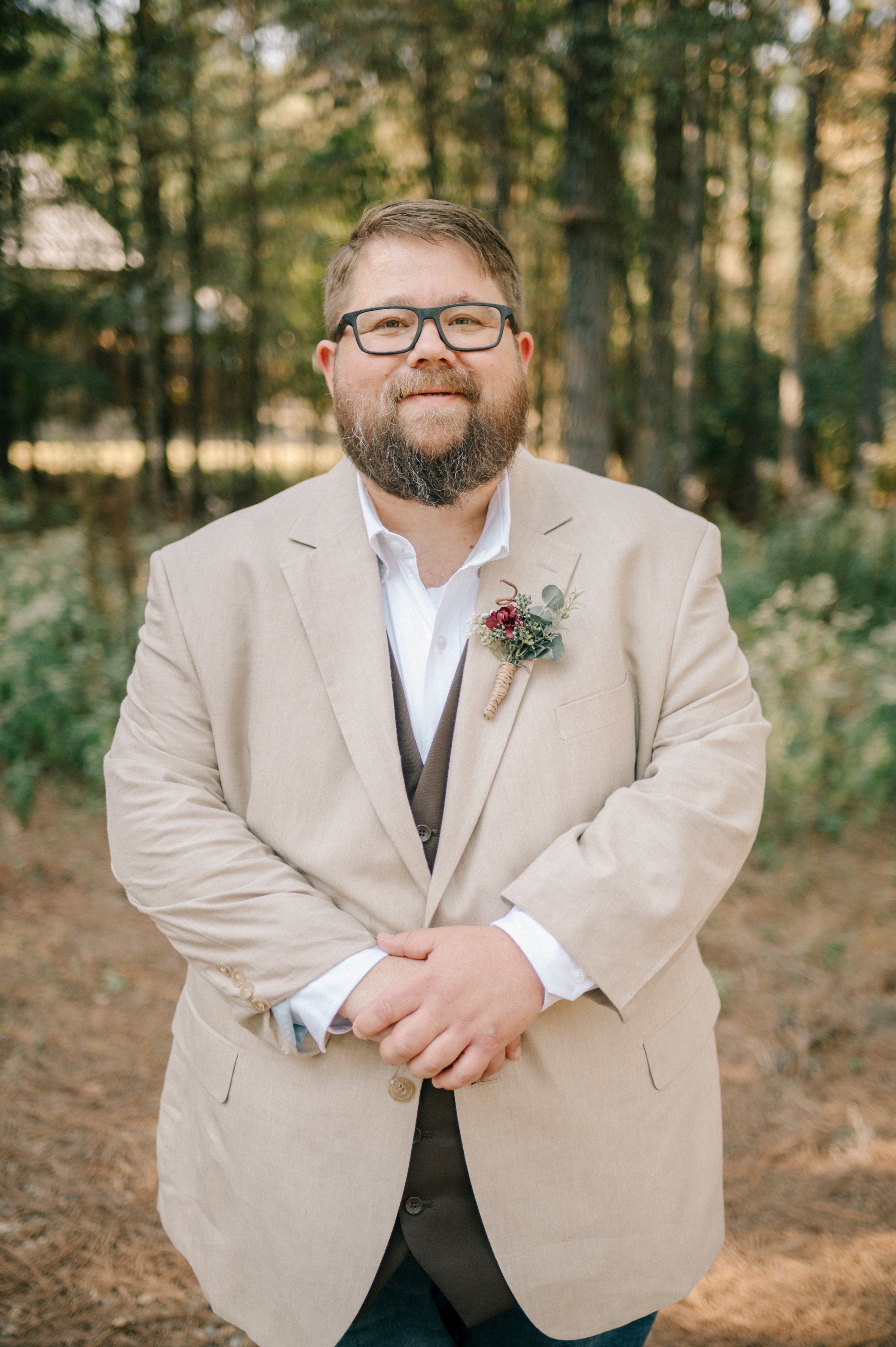 A man with glasses and a beard, dressed in a beige suit with a white shirt, standing outdoors in a wooded area with sunlight filtering through trees. Jacob Caldwell of Ironclad Surfaces, a professional concrete coatings expert in Lubbock, Texas.