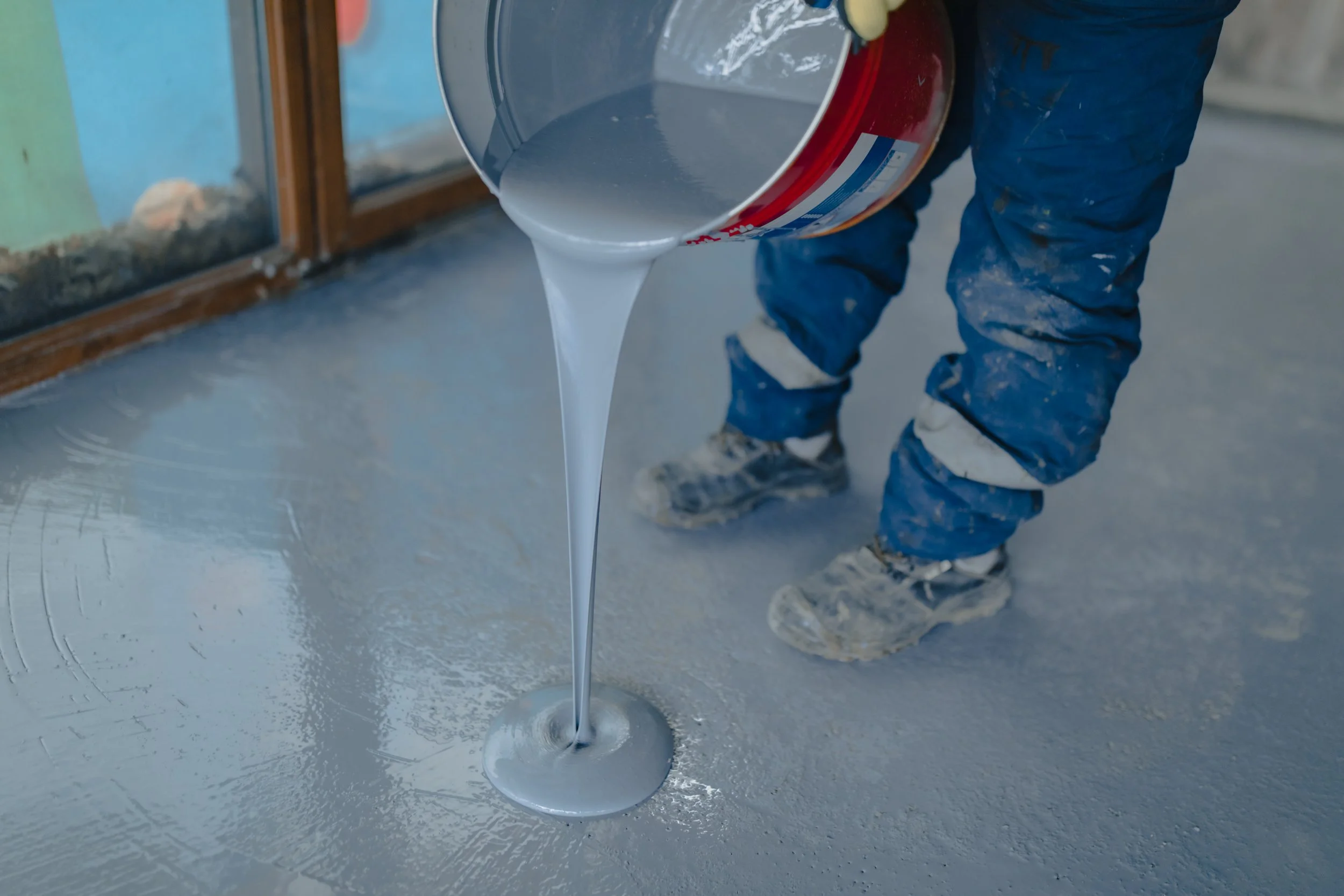 Person pouring liquid waterproofing material onto a surface, with legs in protective clothing visible.