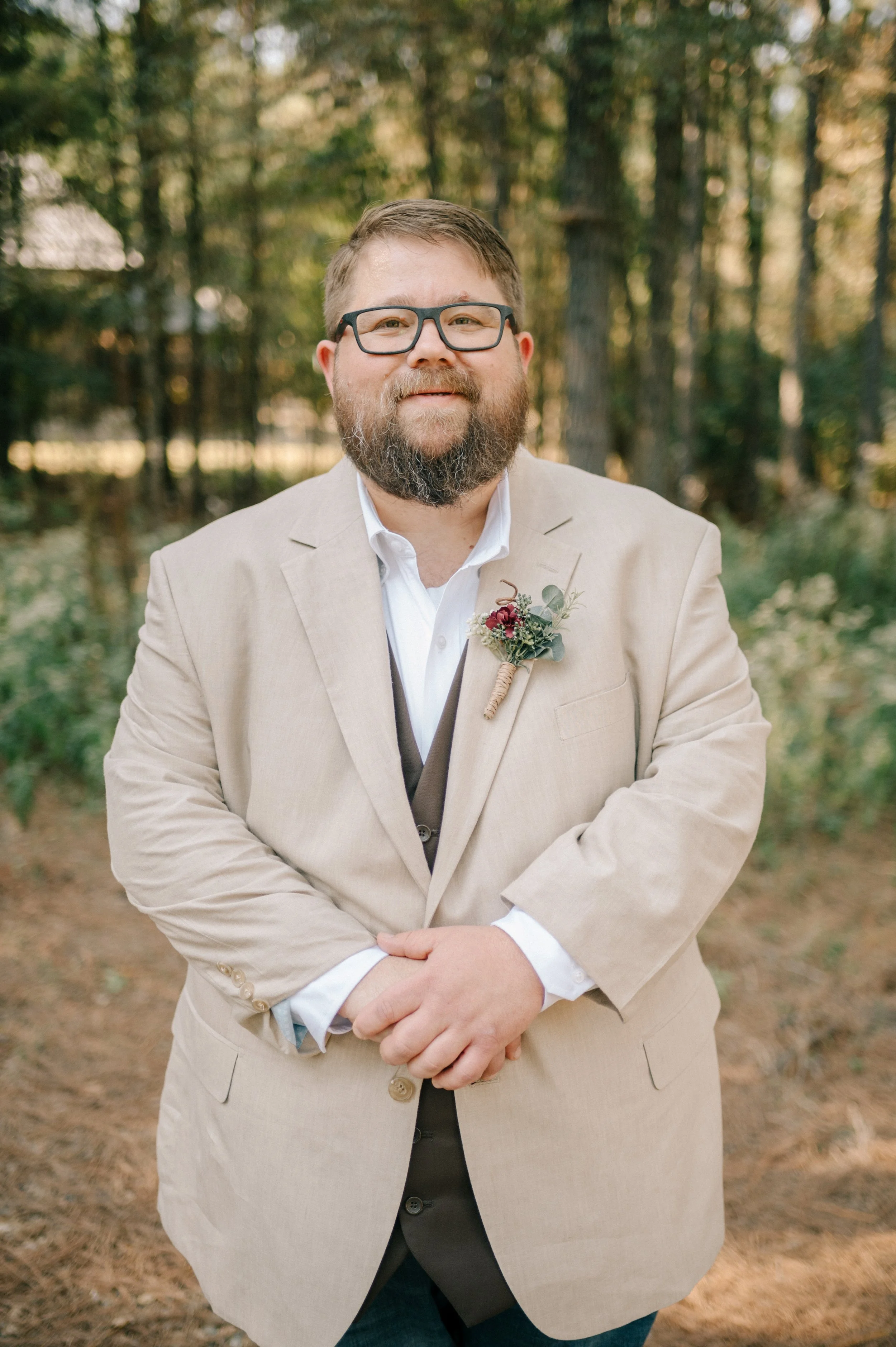A man with glasses and a beard in a beige suit jacket and white shirt standing outdoors in a wooded area, with a boutonniere pinned to his lapel.